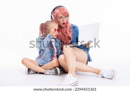 Happy mother and preschooler daughter sitting on white background in studio isolated posing for camera in denim clothes watching video cartoons on laptop. Mom in pink wig and daughter with braids.