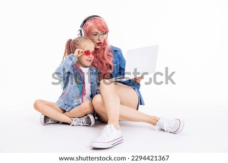 Mother in pink wig with headphones wearing glasses looking at laptop screen watching cartoons with pretty daughter with braids sitting on white floor in studio isolated posing for camera.