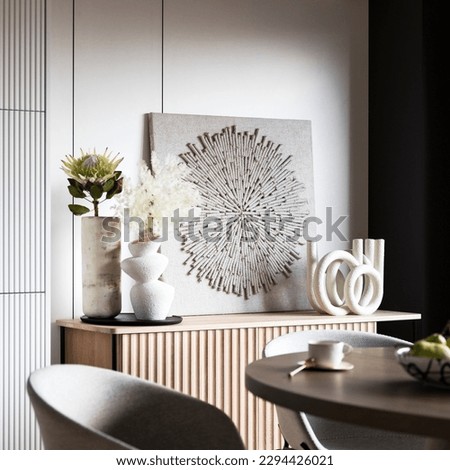 Interior design of aesthetic and elegant room with stylish commode, round table, grey chair, flowers in modern vases, beautiful paintings, candlestick and elegant personal accessories. Home decor.
