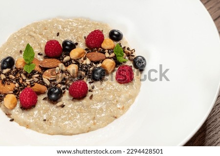 Oatmeal with nuts of various kinds, raspberries and blueberries with mint leaves in a light ceramic plate. The dish stands on a wooden table, next to a glass with orange juice.