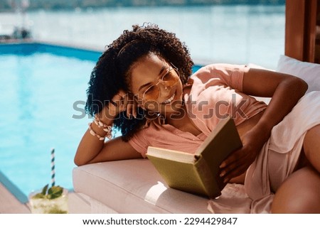Happy African American woman enjoying on summer vacation while reading novel by swimming pool. Royalty-Free Stock Photo #2294429847