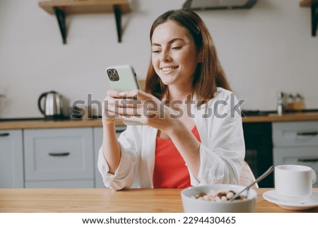 Young fun housewife woman wear casual clothes hold in hand use mobile cell phone eat breakfast muesli cereals with milk fruit in bowl cooking food in light kitchen at home alone. Healthy diet concept