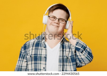 Young smiling satisfied man with down syndrome wear glasses casual clothes headphones listen to music look camera isolated on pastel plain yellow color background. Genetic disease world day concept