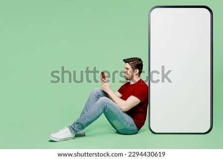 Full body side view young man he wears red t-shirt casual clothes sit near big huge blank screen mobile cell phone with workspace area using smartphone isolated on plain pastel light green background Royalty-Free Stock Photo #2294430619