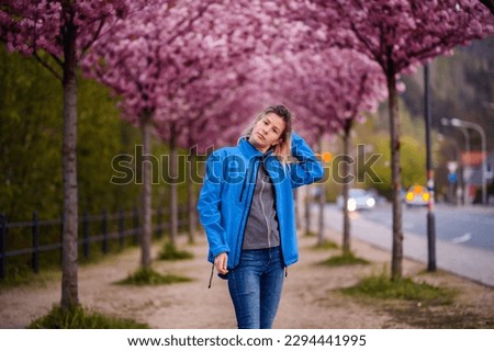 Yong blonde woman in blue jacket and jeans stands in pink blooming sacura alley