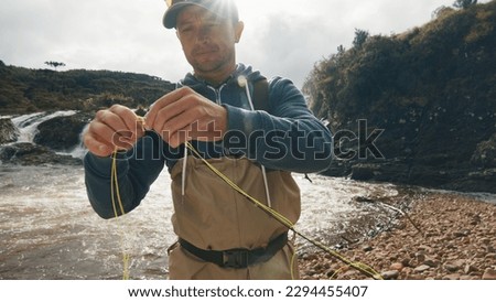 Angler prepares for fly fishing. Fisherman in waders stands by the rapid murky river and prepares the tackle for fishing Royalty-Free Stock Photo #2294455407