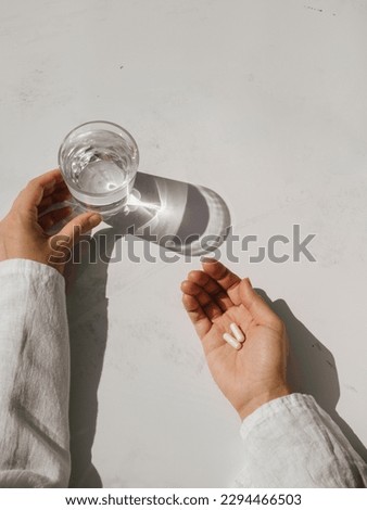 Two capsules on a woman's palm and a glass of water holding by her hand on a white and grey background in sunlight. Top view. Copy space. Royalty-Free Stock Photo #2294466503