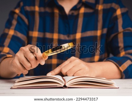 Close-up of hand holding a magnifying glass and reading a book while sitting at the table. Education and learning concept