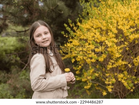 Beautiful girl in a beige raincoat with a yellow dandelion in her hands in a blooming spring park. Girl 8 years old in spring on the background of dandelions, yellow bushes, flower and grass.
