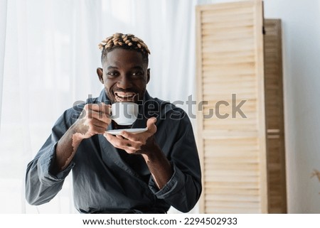 Happy african american man with vitiligo holding coffee and looking at camera at home