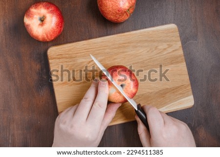 The man is cutting red apple on wooden chopping board. The flat lay of cutting board with red apples on the brown table. The dark food photography of apples and cook hands with knife.