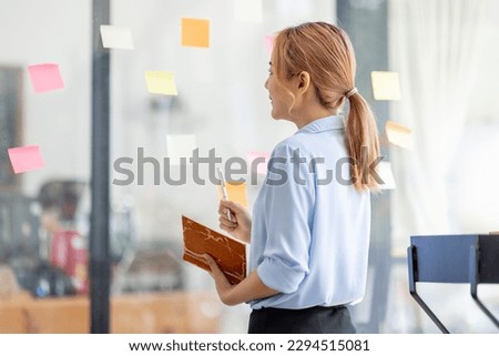 Young business asian woman creative team using post it notes in glass wall to writing finance expert strategy business plan to development grow to success.