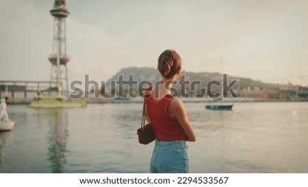 Girl stands on the embankment and looks at the bay. Young woman looks at the sea in the morning time. Back view Royalty-Free Stock Photo #2294533567