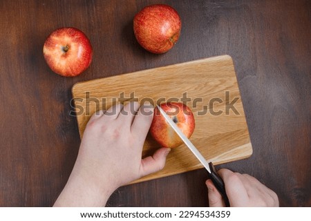 Man is cutting a red apple on wooden chopping board. The flat lay of cutting board with red apples on the brown table. The dark food photography of apples and cook hands with knife.