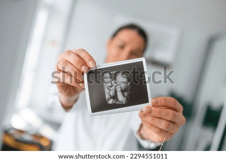 Standing and smiling. Woman in white coat is holding ultrasound picture of baby photo.
