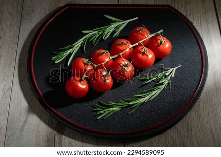 A black tray with branches of cherry tomatoes and Rosmarinus is on a wooden table. It is a dark food photography of red tomatoes and Rosmarinus.