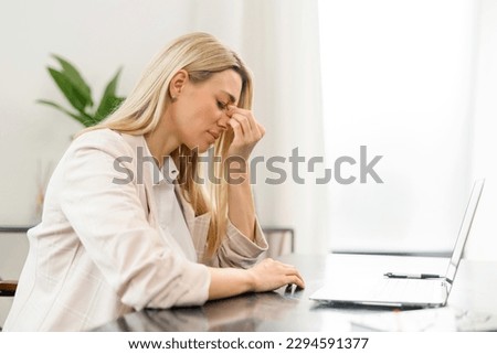 Tired female feeling bored and tired. Attractive blonde woman sitting with eyes closed in front of the laptop, feeling headache and burnout, holding bridge of yhe nose with eyes closed