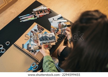 Image from above of an unrecognizable middle-aged woman selecting photos to be taped with washi tape in her handmade kraft travel album. Royalty-Free Stock Photo #2294599455
