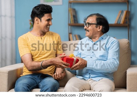 Happy smiling indian adult son giving surprise gift or present to father while sitting on sofa at home - concept of father's day, joyful moment and bonding