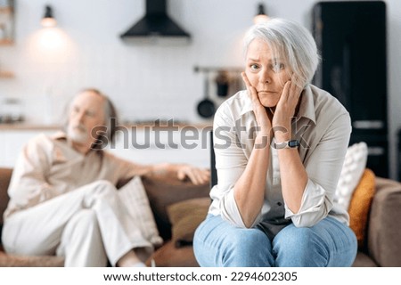 Senior caucasian spouses misunderstanding each other, experiencing crisis in relationship, offended wife looks disappointedly at camera, husband sits in defocus on background. Quarrel, marital crisis