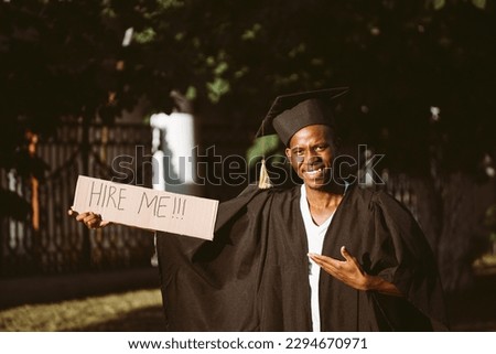 Portrait of smiling black guy standing and pointing at cardboard poster on street in sunny day looking for job, employment issue. University or college graduating student in graduate gown and cap. Royalty-Free Stock Photo #2294670971
