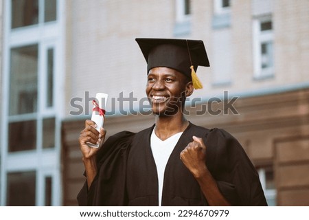 Portrait of smiling black guy graduate from university standing outdoors near building and holding in hand higher education diploma, selective focus. Graduation mantle and hat, ceremonial celebration. Royalty-Free Stock Photo #2294670979