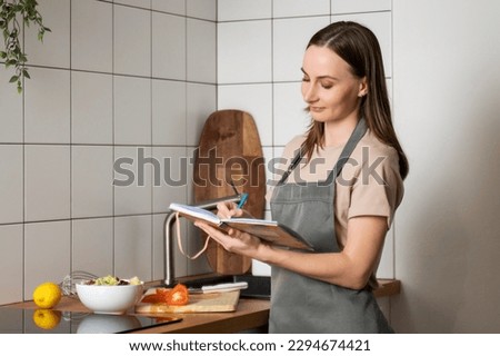 A smiling young beautiful business woman makes notes in a paper diary in a modern kitchen. A happy woman writing in a notebook surrounded by a convenient storage organization. 