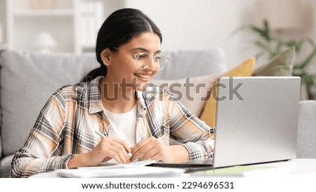 A young Indian girl works at a laptop and looks at the monitor. Beautiful woman talking on video call smiling. Studying in quarantine, listening to a podcast. Royalty-Free Stock Photo #2294696531