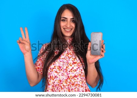 beautiful brunette woman wearing floral dress over blue background holding modern device showing v-sign