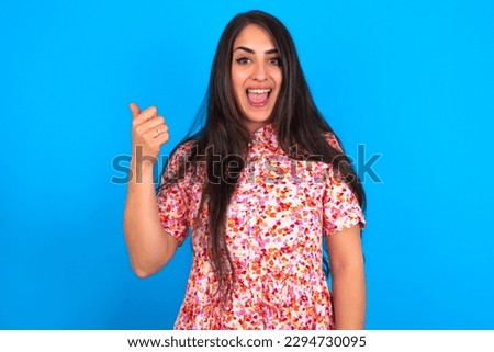 Impressed beautiful brunette woman wearing floral dress over blue background point back empty space