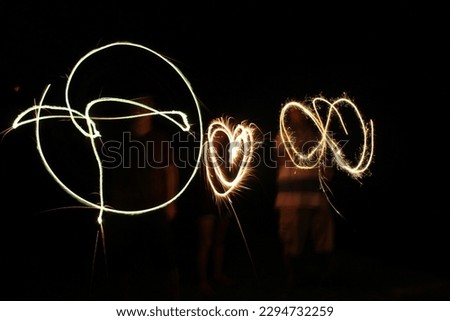 Three people stand in the pitch black as they move firework sparklers as the camera captures it with a slow shutter speed.