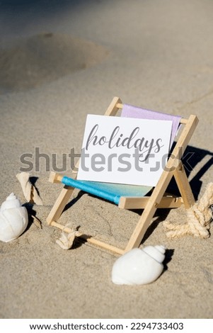 HOLIDAYS text on paper greeting card on background of beach chair lounge summer vacation decor. Sandy beach sun. Holiday concept postcard. Travel