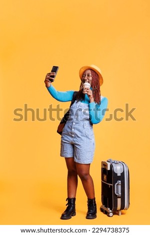Young adult taking photos on vacation, drinking cup of latte on camera. Female traveller with luggage bag feeling happy on international holiday, enjoying urban adventure with images.