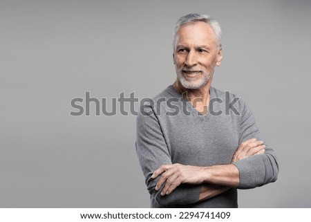Emotional studio portrait of handsome confident elderly senior gray haired man in casual clothes, posing with his arms crosse, smiling looking away at copy advertising space, isolated grey background Royalty-Free Stock Photo #2294741409