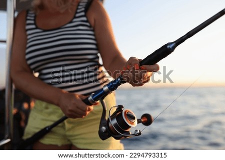 Woman in striped t-shirt enjoys fishing with rod on boat. Female person tries to catch fresh fish in sea at bright sunset on clear sky Royalty-Free Stock Photo #2294793315