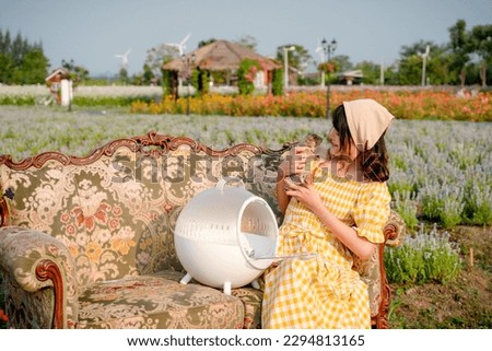 Cheerful pretty young woman with hair tied by handkerchief holding and hugging cute little prairie dog sitting on vintage sofa relaxing in beautiful flower garden. Asian people.