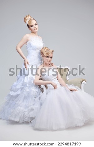 Full-length portrait of two blonde ladies posing together in white wedding dresses with avant-garde hairstyles and make-up. Studio shot on a gray background. Wedding Fashion. 