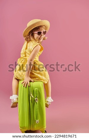 A little funny girl in a bright yellow summer dress, sunglasses and a hat sits on a suitcase with her back to the camera and laughs. Time to go on vacation!  Pink studio background with space for text