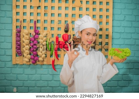Beautiful asian female professional chef holding a bowl of fresh green vegetables in kitchen full of cooking ingredients with happy expression and make ok hand sign.