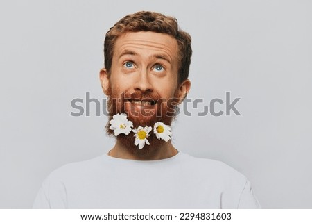 Portrait of a funny man in a white T-shirt with flowers daisies in his beard on a white isolated background, copy place. Holiday concept and congratulations.