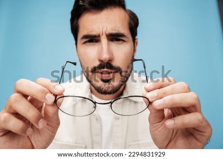 Close-up portrait of a man looking through glasses that he holds in his hands, dioptric lenses, glasses for far-sightedness and near-sightedness, on a blue background in a white T-shirt, copy space 