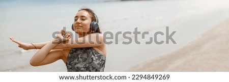 Beautiful young sportive woman in headphone making stretching exercises on beach. Blurred background