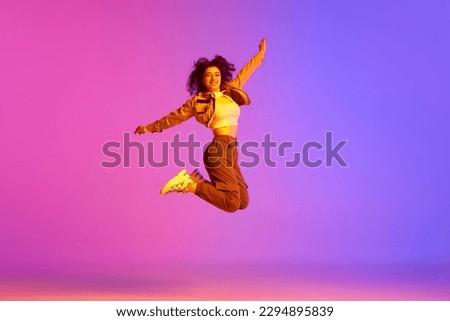 Young girl, hip-hop dancer in sport style clothes jumping against gradient pink purple background in neon light. Delightful. Concept of contemporary dance, youth, hobby, action and motion