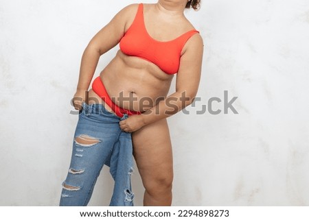 Unrecognizable adipose, fat, obese and overweight woman in red bikini pulling and getting dressed in small size tight jeans, to fit hips in clothes on white background. Preparation summer season