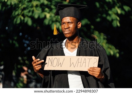 Portrait of discontented  american guy holding and pointed at cardboard poster on street. Looking for job, hiring and employment issue. University or college graduating student in gown and cap. Royalty-Free Stock Photo #2294898307