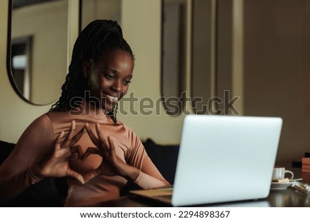 Portrait of young smiling African-American woman with long dark braids wearing pink roll-neck, sitting at table in cafe near laptop, showing heart with fingers, love sign at screen. Communication.  Royalty-Free Stock Photo #2294898367
