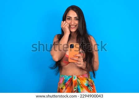 Joyous beautiful brunette woman wearing swimwear over blue background poses with mobile phone device, types text message on modern smartphone, watches funny video during free time, enjoys good interne