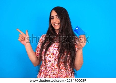 Curious smiling beautiful brunette woman wearing floral dress over blue background showing plastic bank showing finger copyspace