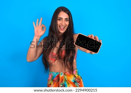 beautiful brunette woman wearing swimwear over blue background holding in hands cell showing ok-sign
