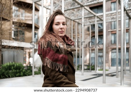 Woman looking at camera with metal structure behind. She is between buildings and she is wearing coat and scarf.